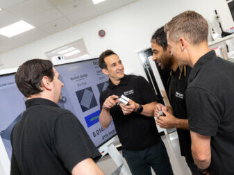 Ricoh 3D's Head of Additive Manufacturing, Mark Dickin, holding a 3D-printed part and conversing with the engineering team in front of a large digital monitor.